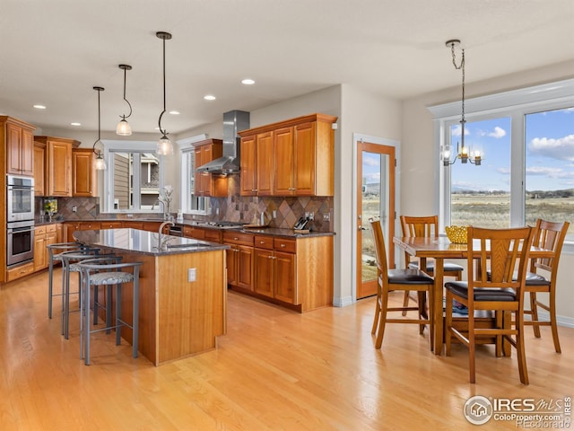 kitchen with brown cabinets, a center island with sink, stainless steel appliances, light wood-style floors, and wall chimney exhaust hood