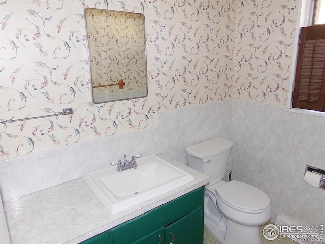 half bathroom featuring vanity, wallpapered walls, wainscoting, tile walls, and toilet