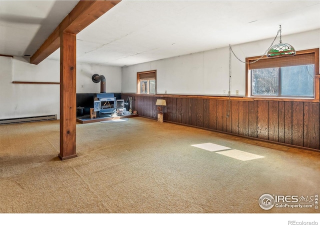 basement featuring wooden walls, a wainscoted wall, carpet flooring, a wood stove, and a baseboard radiator