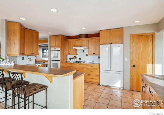 kitchen with under cabinet range hood, a breakfast bar, light tile patterned floors, decorative backsplash, and white appliances