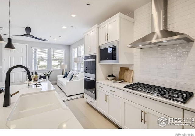 kitchen featuring a sink, stainless steel appliances, white cabinets, wall chimney range hood, and hanging light fixtures