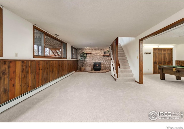 interior space featuring a wood stove, a baseboard heating unit, carpet floors, wood walls, and stairs