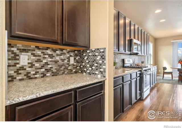 kitchen featuring dark brown cabinets, light stone countertops, decorative backsplash, wood finished floors, and stainless steel appliances