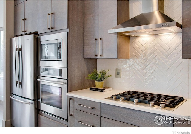 kitchen with gray cabinets, light countertops, appliances with stainless steel finishes, wall chimney exhaust hood, and backsplash