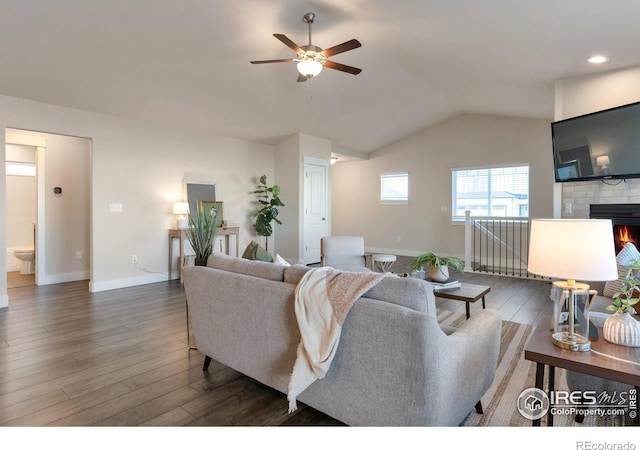 living room featuring a warm lit fireplace, wood finished floors, baseboards, ceiling fan, and vaulted ceiling