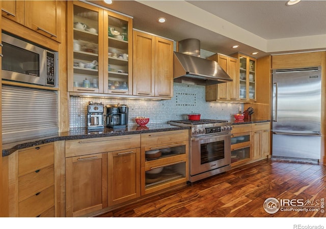 kitchen with dark wood finished floors, glass insert cabinets, built in appliances, wall chimney range hood, and tasteful backsplash