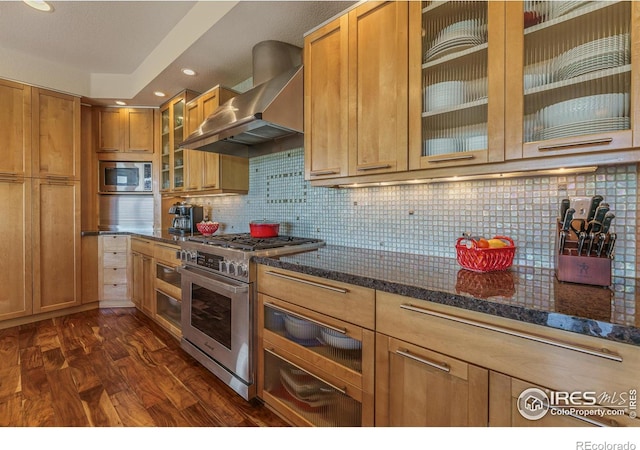 kitchen featuring dark stone countertops, dark wood-style flooring, stainless steel appliances, wall chimney exhaust hood, and backsplash