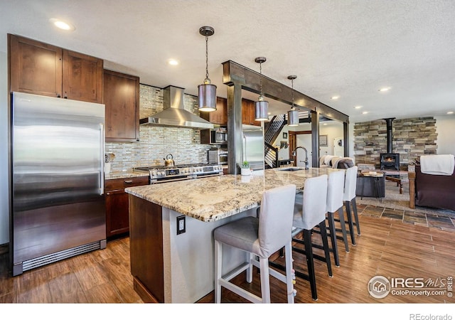 kitchen featuring wood finished floors, stainless steel appliances, wall chimney exhaust hood, decorative backsplash, and a wood stove