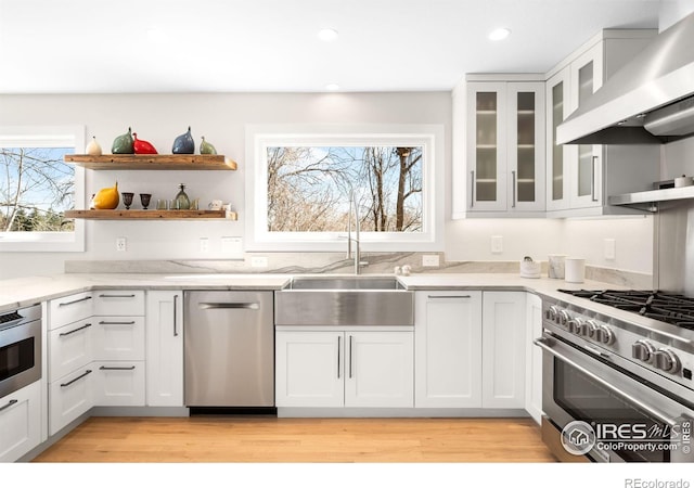 kitchen featuring open shelves, light stone counters, appliances with stainless steel finishes, wall chimney exhaust hood, and a sink