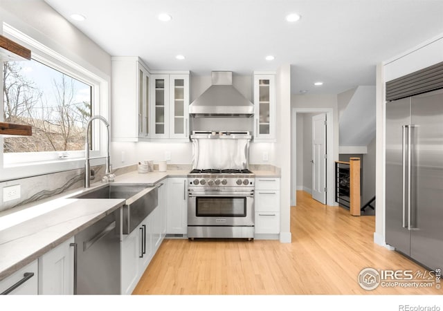 kitchen with a sink, light stone counters, white cabinetry, wall chimney exhaust hood, and high end appliances