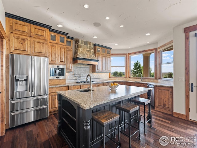 kitchen featuring a center island with sink, dark wood finished floors, a sink, custom range hood, and appliances with stainless steel finishes