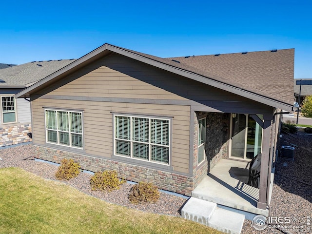 rear view of property with central AC unit, a patio area, stone siding, and a shingled roof