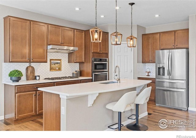 kitchen featuring brown cabinets, a kitchen island with sink, under cabinet range hood, a sink, and stainless steel appliances