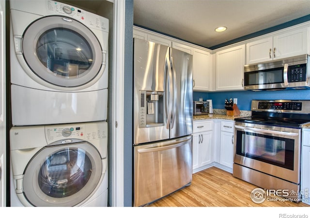 kitchen with light wood-type flooring, light stone counters, appliances with stainless steel finishes, white cabinets, and stacked washer / drying machine