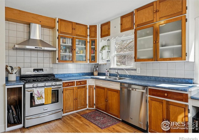 kitchen featuring backsplash, wall chimney range hood, light wood-type flooring, brown cabinetry, and stainless steel appliances