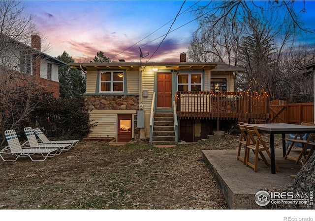 back of property at dusk with a deck, fence, stone siding, and a chimney