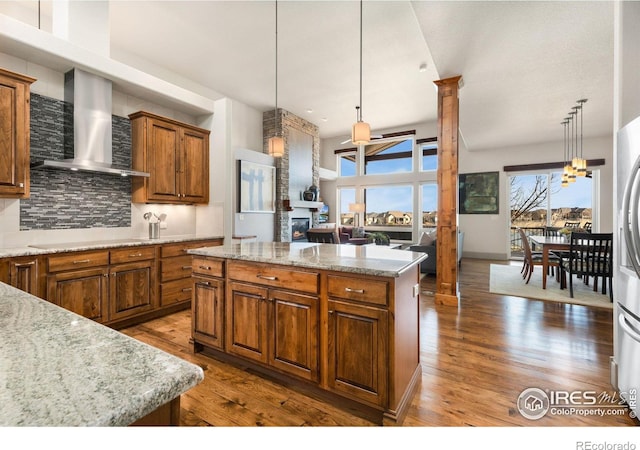 kitchen with wood finished floors, brown cabinets, decorative columns, wall chimney exhaust hood, and tasteful backsplash