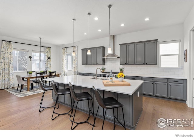 kitchen featuring an island with sink, light wood-style flooring, gray cabinets, and wall chimney range hood