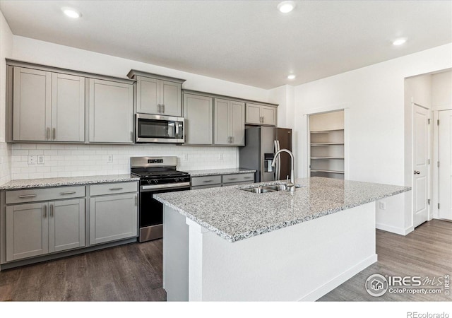 kitchen with gray cabinetry, an island with sink, a sink, appliances with stainless steel finishes, and dark wood-style flooring