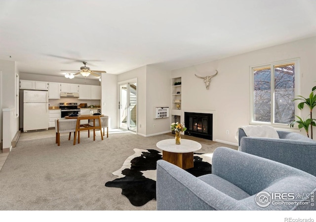 living room with built in shelves, a glass covered fireplace, baseboards, light colored carpet, and ceiling fan