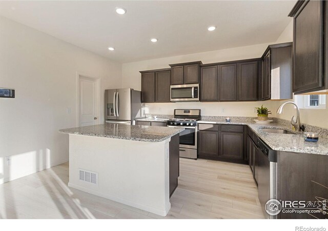 kitchen featuring light stone counters, a sink, stainless steel appliances, dark brown cabinetry, and a center island