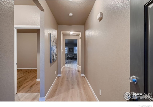 corridor featuring light wood-type flooring, baseboards, a textured ceiling, and a textured wall