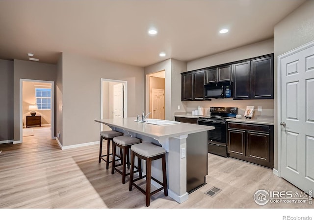 kitchen featuring black appliances, a center island with sink, a sink, light wood-style floors, and light countertops