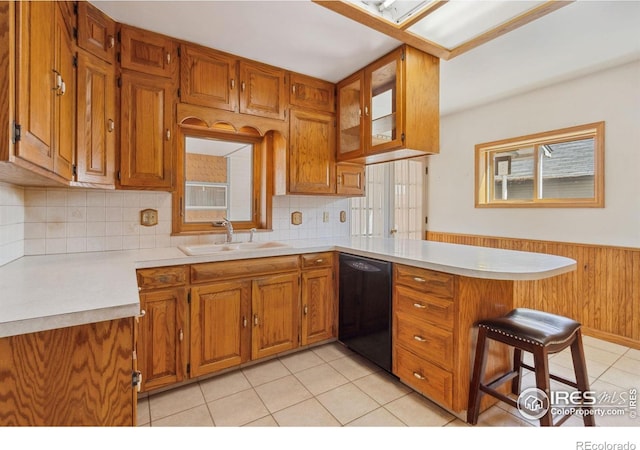 kitchen with black dishwasher, a peninsula, brown cabinetry, wainscoting, and light countertops