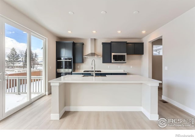 kitchen featuring baseboards, an island with sink, stainless steel appliances, light countertops, and wall chimney range hood