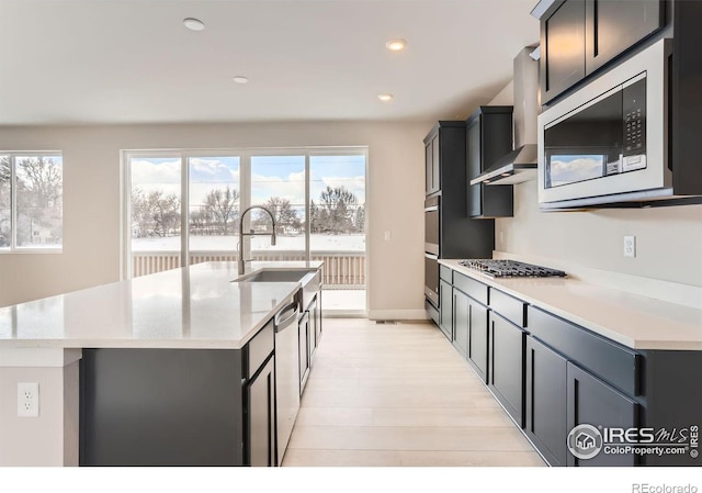 kitchen featuring recessed lighting, a kitchen island with sink, stainless steel appliances, a sink, and wall chimney range hood