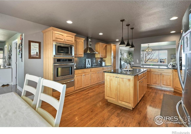 kitchen featuring light brown cabinetry, wall chimney range hood, wood finished floors, dark countertops, and stainless steel appliances