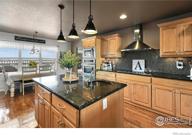 kitchen featuring an island with sink, light wood-type flooring, wall chimney range hood, a sink, and stainless steel appliances