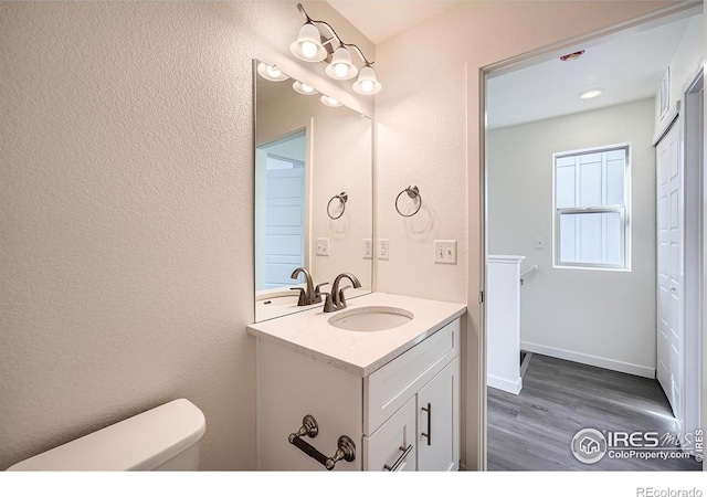 half bathroom with toilet, wood finished floors, baseboards, vanity, and a textured wall