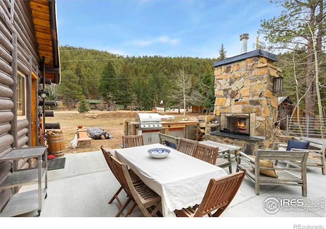 view of patio featuring a grill, outdoor dining area, an outdoor stone fireplace, and a wooded view