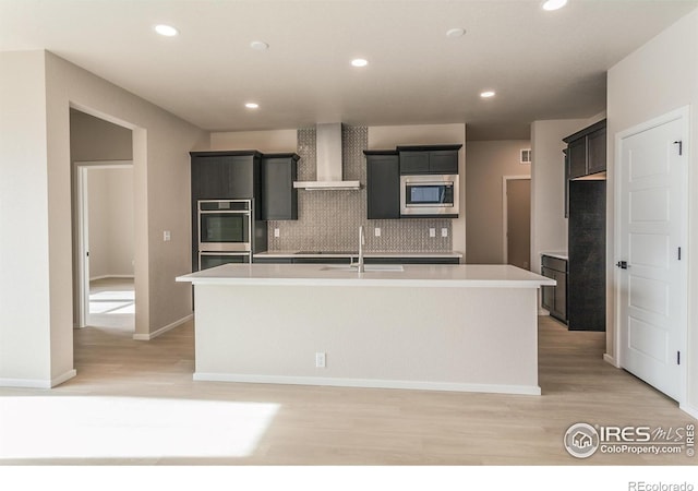 kitchen featuring stainless steel microwave, an island with sink, decorative backsplash, wall chimney exhaust hood, and a sink