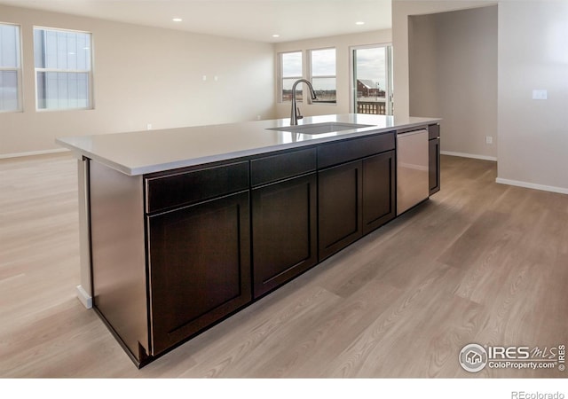 kitchen with light wood-style flooring, an island with sink, a sink, stainless steel dishwasher, and light countertops