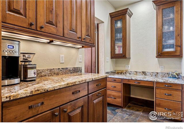 kitchen with brown cabinets, stone finish flooring, glass insert cabinets, light stone countertops, and built in study area