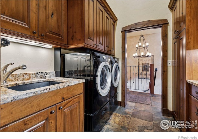 clothes washing area with washing machine and clothes dryer, a chandelier, stone tile floors, cabinet space, and a sink