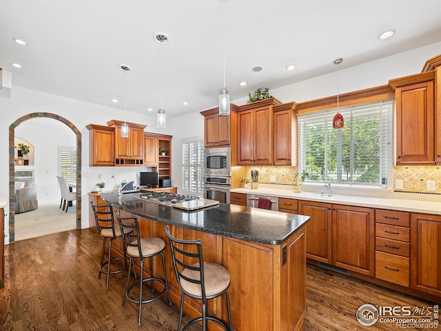 kitchen with dark wood finished floors, a kitchen island, arched walkways, appliances with stainless steel finishes, and brown cabinets