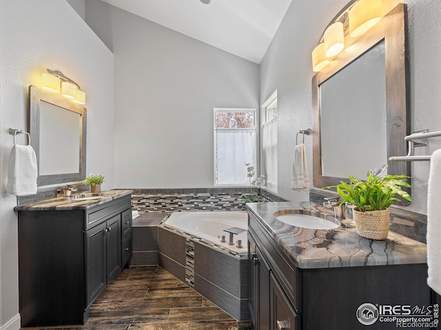 bathroom with a bath, wood finish floors, two vanities, a sink, and vaulted ceiling