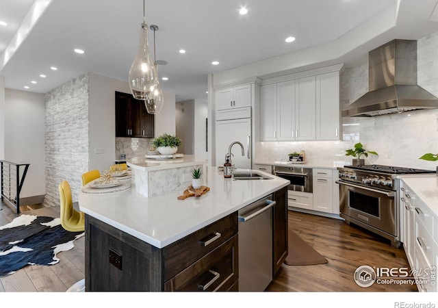 kitchen featuring high end appliances, a sink, dark wood-type flooring, wall chimney range hood, and a kitchen island with sink