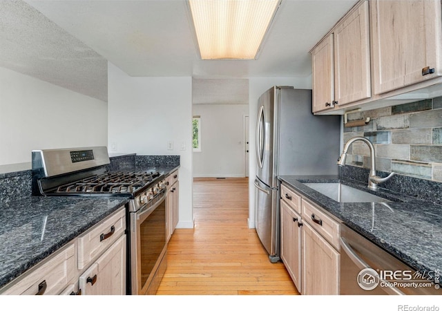 kitchen featuring light brown cabinets, light wood-style flooring, dark stone counters, appliances with stainless steel finishes, and a sink