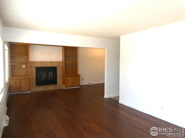 unfurnished living room with a tiled fireplace, dark wood-type flooring, visible vents, and baseboards