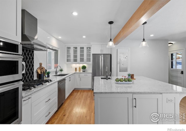 kitchen with white cabinetry, stainless steel appliances, a sink, a kitchen island with sink, and wall chimney exhaust hood