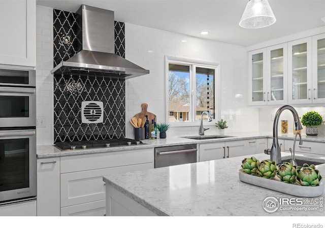 kitchen featuring appliances with stainless steel finishes, white cabinetry, a sink, wall chimney range hood, and backsplash