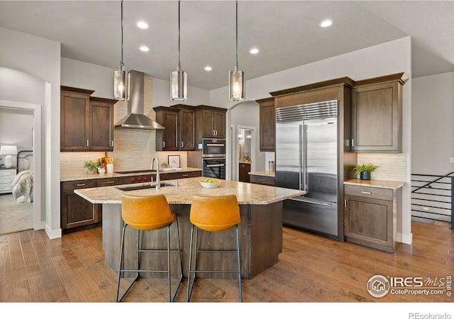 5977 Crooked Stick Dr, Windsor, CO 80550 - kitchen with built in refrigerator, wall chimney exhaust hood, wood finished floors, an island with sink, and backsplash