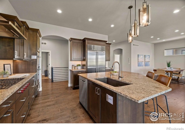 5977 Crooked Stick Dr, Windsor, CO 80550 - kitchen with stainless steel appliances, arched walkways, wall chimney range hood, a kitchen island with sink, and dark wood-type flooring