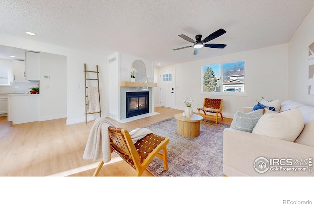living area with ceiling fan, a fireplace with flush hearth, a textured ceiling, light wood-type flooring, and baseboards