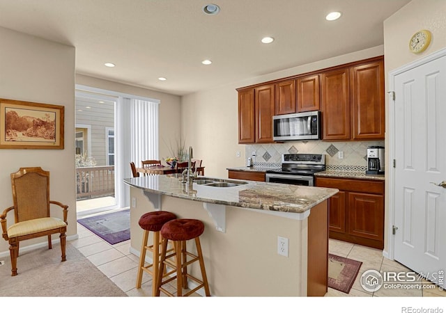 kitchen with light tile patterned floors, appliances with stainless steel finishes, light stone counters, and an island with sink
