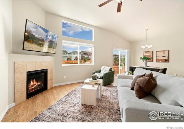 living room with ceiling fan with notable chandelier, vaulted ceiling, wood finished floors, a tiled fireplace, and baseboards